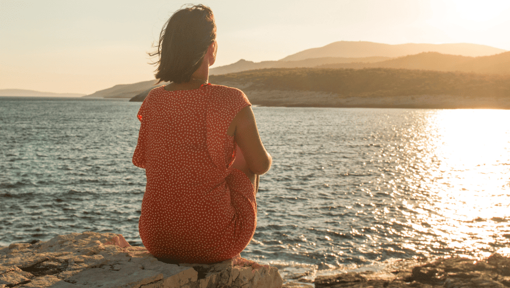 respirar, refletir e agradecer, mulher sentada na beirada de uma pedra de frente para o mar, observando o nascer do sol.