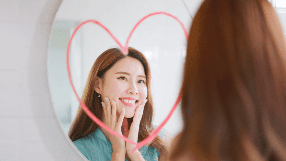 Frases para Aumentar Autoestima 200 Maneiras de Se Amar Mais Todos os Dias, imagem de uma mulher se olhando no espelho, sorrindo e com um coração desenhado no espelho, simbolizando o amor próprio.