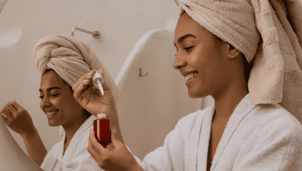 Quais são os 4 tipos de autocuidado - Como iniciar e criar uma rotina de autocuidado - imagem de uma mulher já com banho tomado, com roupão, cabelo lavado, com a toalha na cabeça, em frente ao espelho, se cuidando e aplicando um sérum no rosto.