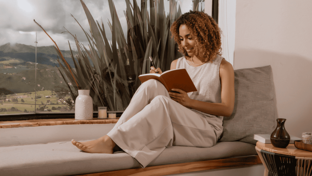 Quais são os 4 tipos de autocuidado - Exemplos práticos de autocuidado - imagem de mulher sentada no sofá ao lado de uma janela transparente com uma paisagem linda, com um caderno anotando todas informações.