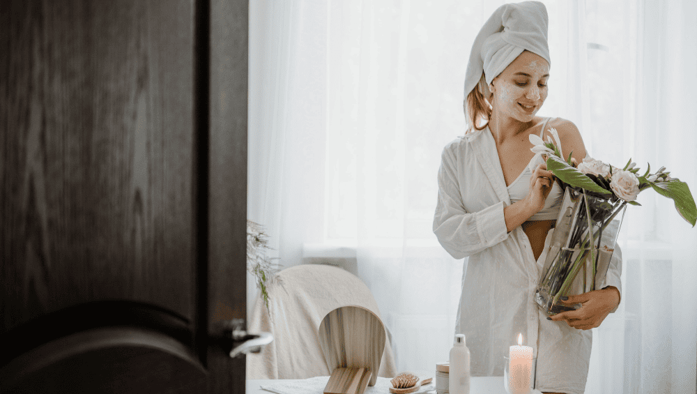 Quais são os 4 tipos de autocuidado - obstáculos comuns e desafios - mulher com um vaso com flores, produtos de autocuidado na mesa e uma vela para representar que o autocuidado não precis ser dificil e que podemos eliminar os desafios do dia a dia na hora de realizar o autocuidado.