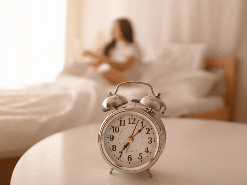 Como Ter Um Dia Produtivo - Comece Seu Dia Com Clareza e Intenção Como Ter Um Dia Produtivo - Comece Seu Dia Com Clareza e Intenção - mulher sentada na cama, após uma bela noite de sono, desfocada atrás de um relogio destacado na imagem.