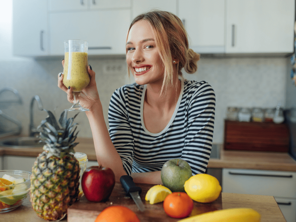 O Que Beber Para Desinflamar o Corpo - Como Incluir Essas Bebidas na Rotina - imagem de uma mulher com um copo na mão com uma bebida saudável, com várias frutas ao redor