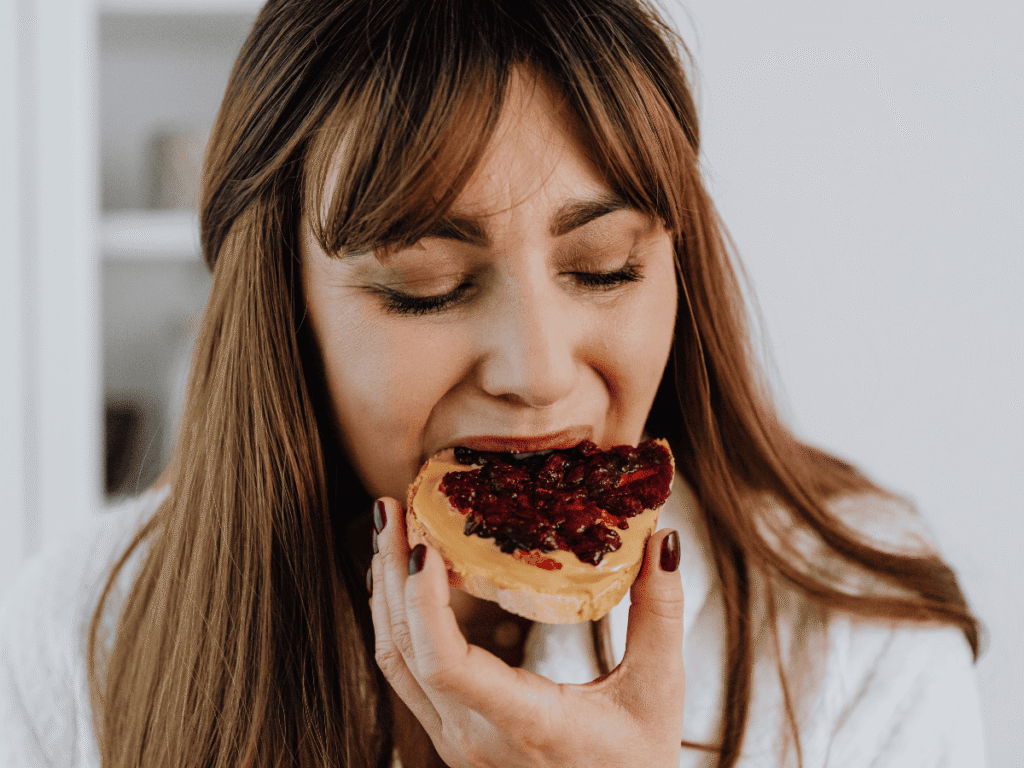 Por Que Você Não Emagrece Nem Comendo Apenas Salada - A Fome Rebote e as Beliscadas Inconscientes - mulher dando uma mordida em uma torrada com doce de leite e geleia.