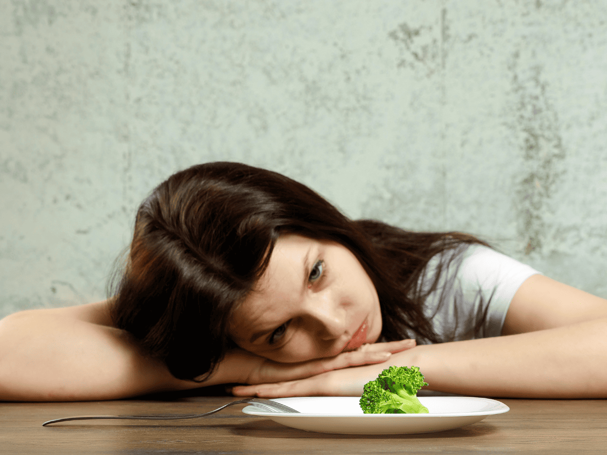 Por Que Você Não Emagrece Nem Comendo Apenas Salada - imagem de uma mulher triste escorada na mesa e um prato de alface na mesa.
